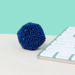 Ein blauer, kugelförmiger Büroklotz mit kleinen Perlen neben einer weißen Tastatur auf einem weißen und mintgrünen Hintergrund.