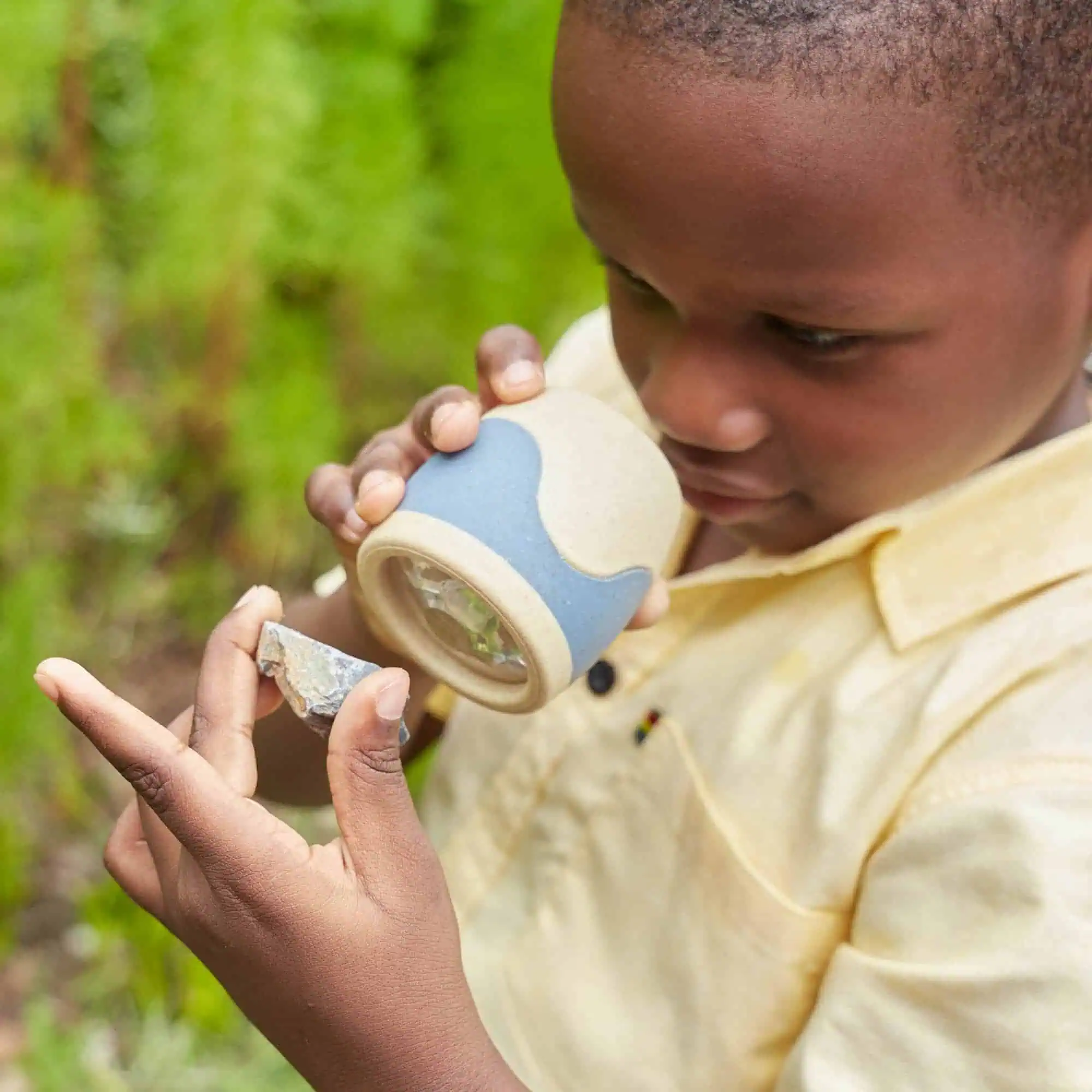 2. Nahaufnahme eines Jungen bei der Untersuchung eines Fossils im Freien, hält eine Lupe und betrachtet vorsichtig ein Fossil mit grünen Pflanzen im Hintergrund.