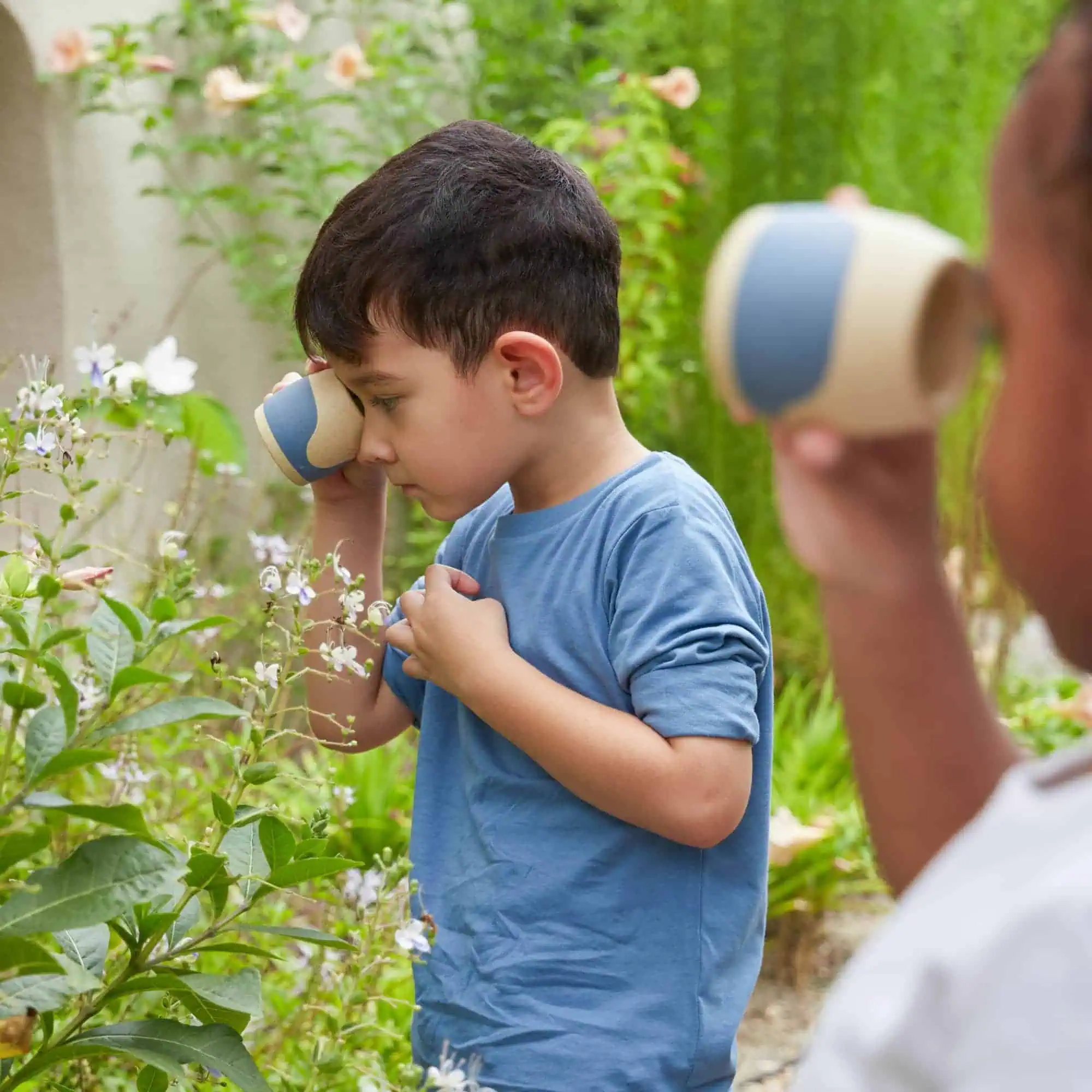 2. Kinder beobachten und riechen an einer Pflanze im Garten mit langen Gräsern und Blüten, während ein Erwachsener mit einem Blumentopf in der Hand zuschaut.