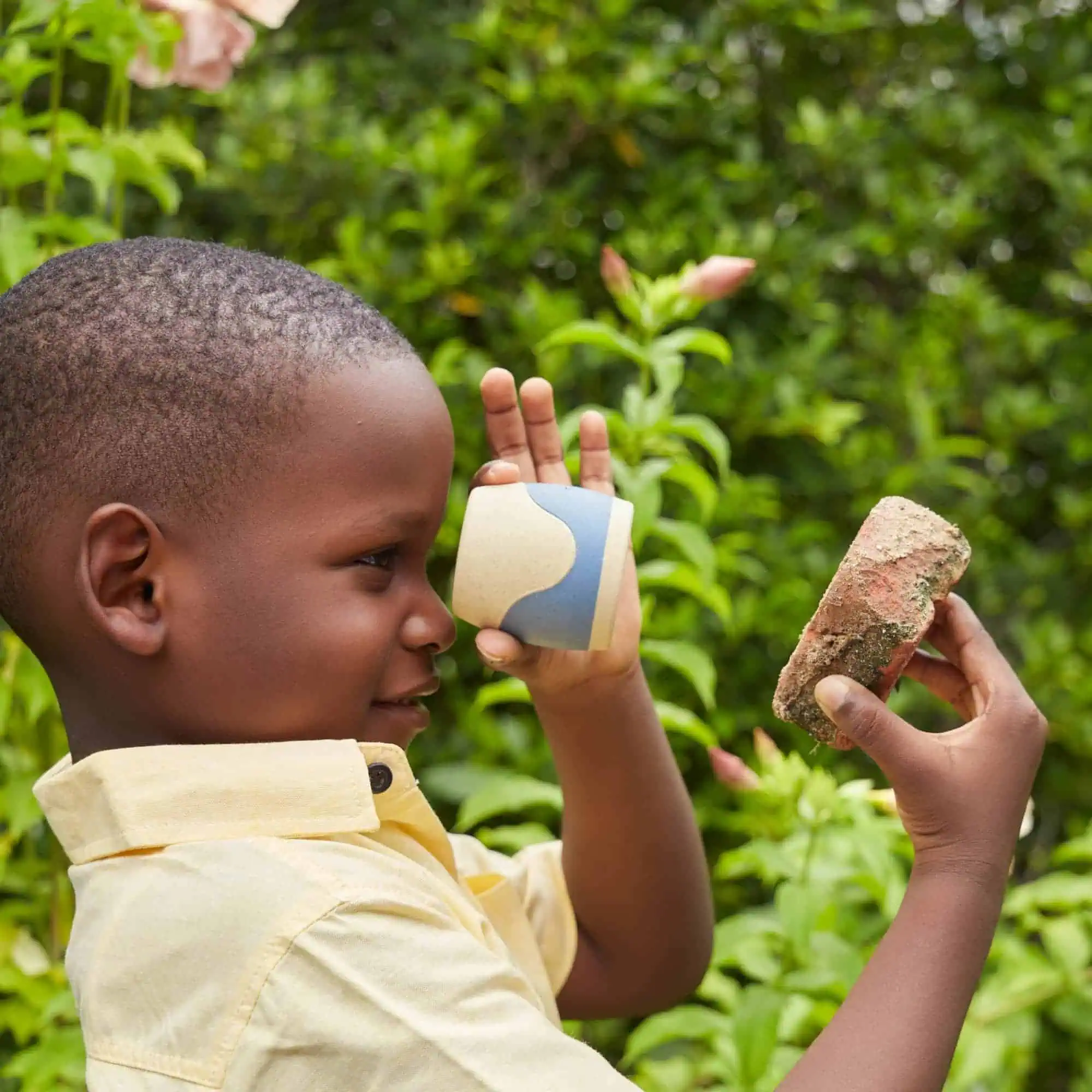 Spielender junger afroamerikanischer junge im garten mit ziegelstein und tasse, grüne blätter im hintergrund.